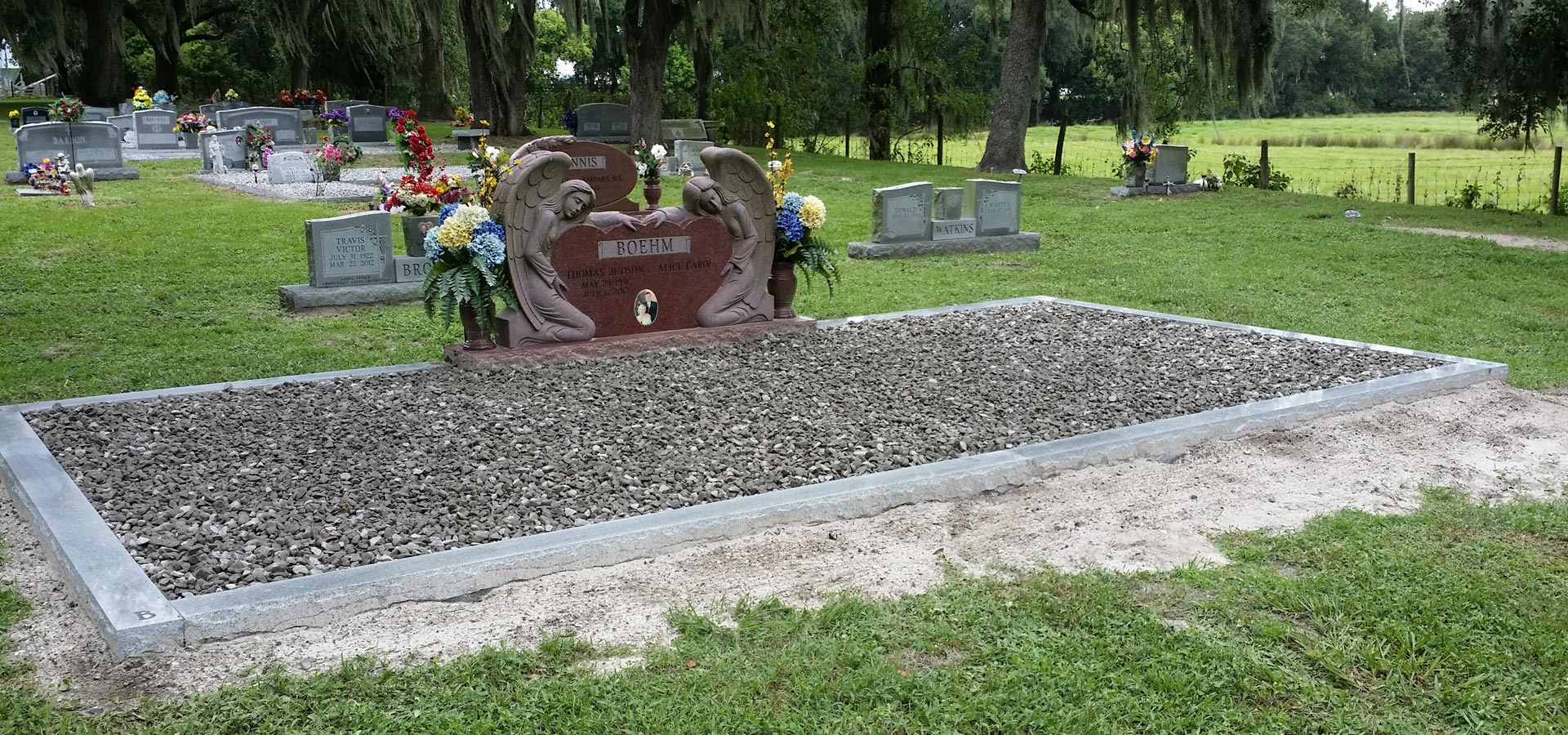 A red granite heart memorial flanked by two carved kneeling angels, standing under live oaks in a Florida cemetery