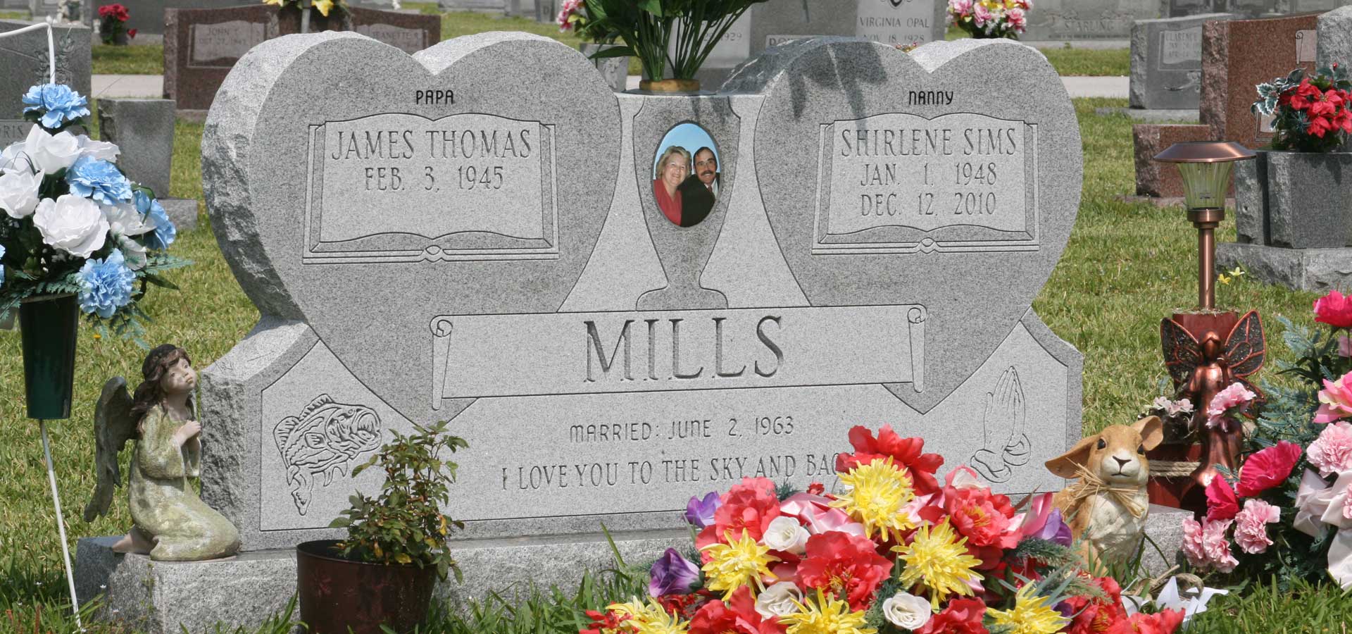 Grey double heart monument with carved open books, a colour portrait and carved detail, surrounded by flowers left by the family